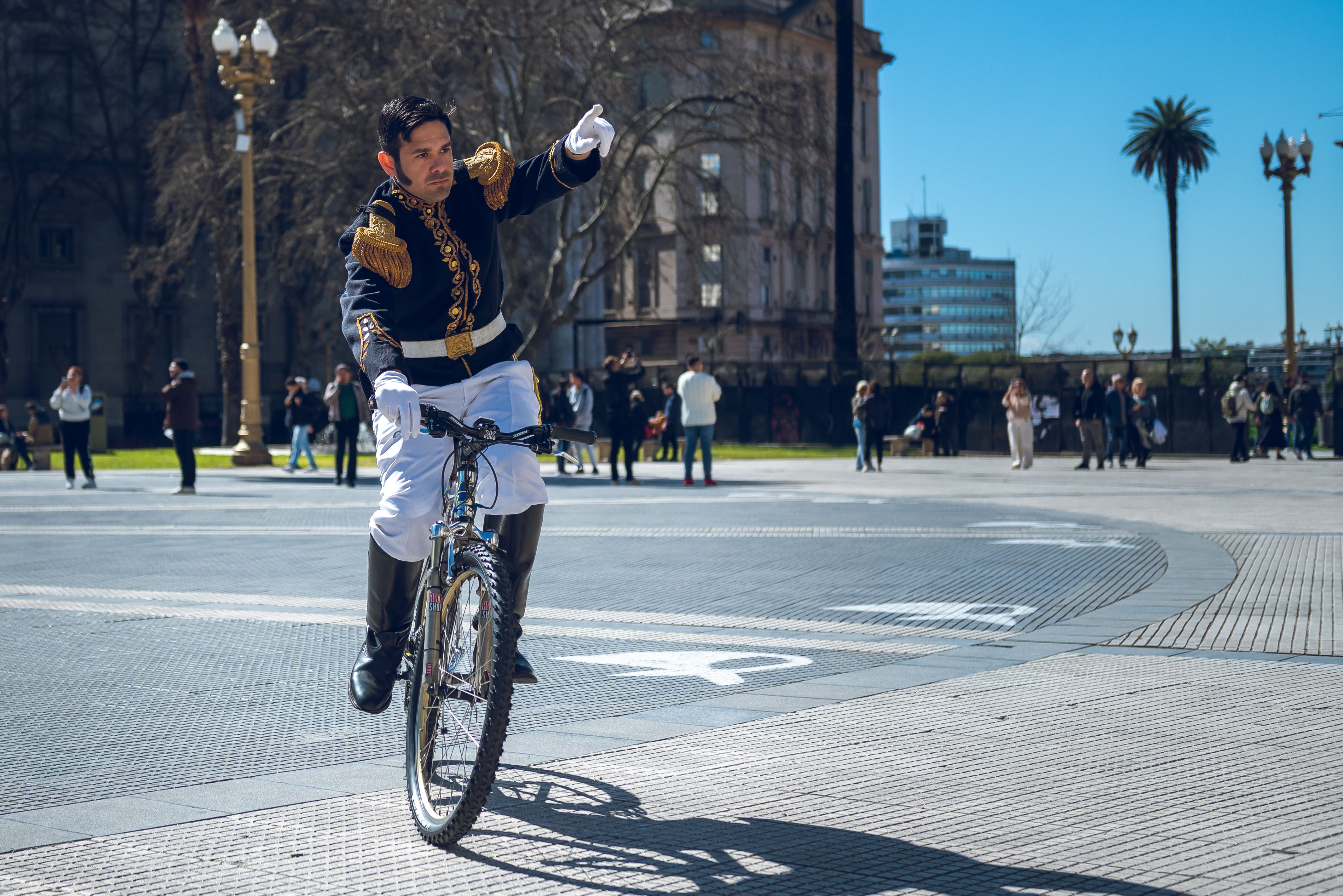 A man in a traditional costume rides a bicycle in Plaza de Mayo, Buenos Aires with people in the background.