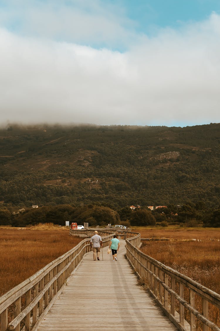 Unrecognizable Senior Couple Walking On Bridge Near Field And Mountains