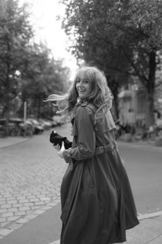 Smiling woman in trench coat walking down a tree-lined street in black and white.