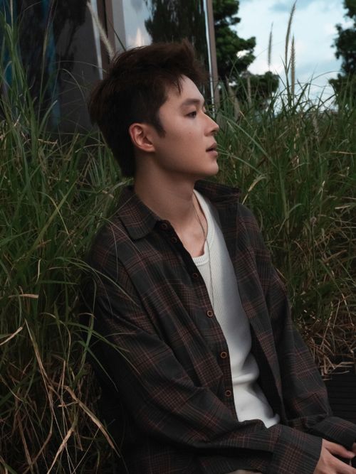 Free Young adult male in plaid shirt sitting thoughtfully in grassy outdoor setting. Stock Photo