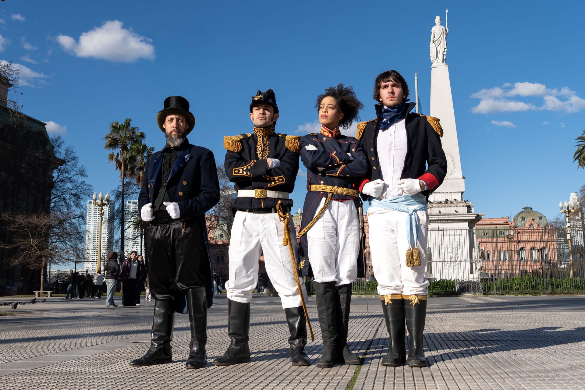 Actors in historical costumes pose near the Obelisco de Buenos Aires. Clear blue skies highlight the scene.