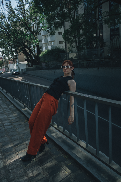 Free Fashionable woman in city wearing red pants and sunglasses, leaning on railing. Stock Photo