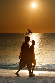 Two silhouettes walking along the beach at sunset with a distant sailboat on the horizon.