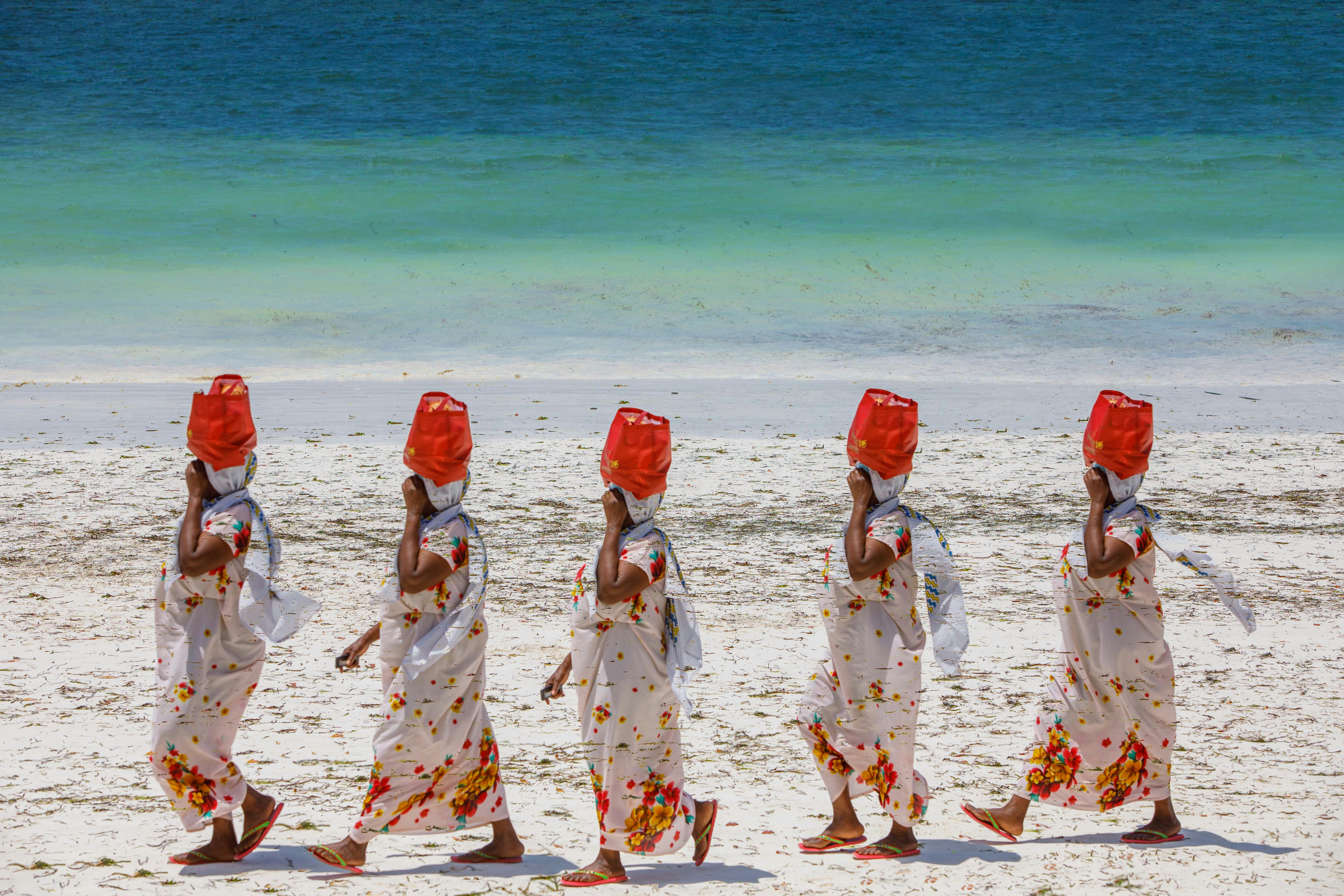 Five women in traditional attire with vibrant scarves walking on a sandy beach by the ocean.