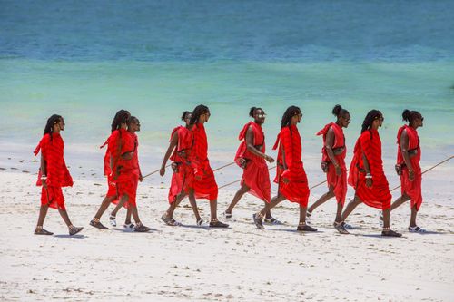 Free Maasai warriors in traditional attire walking along a pristine tropical beach with turquoise waters. Stock Photo