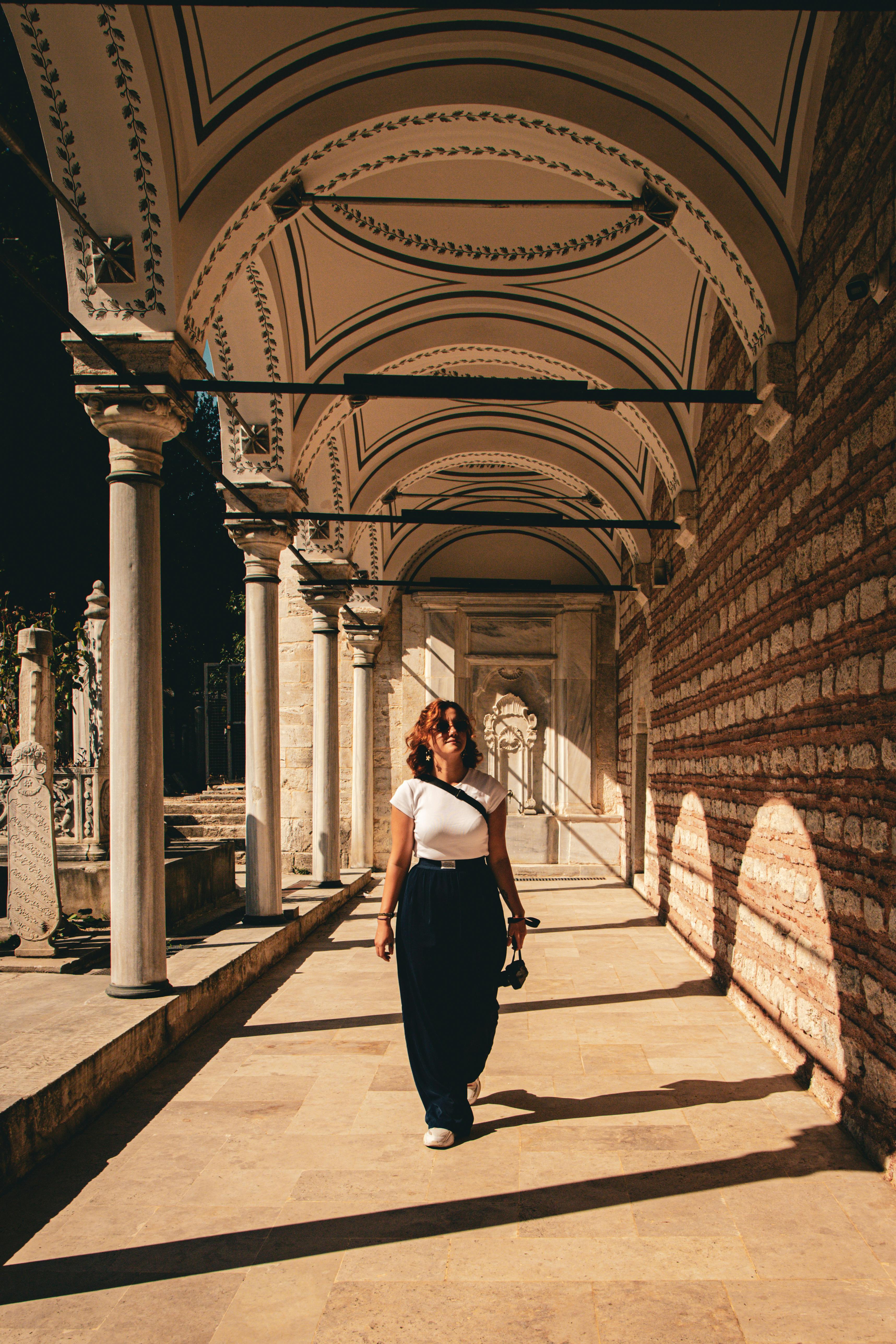 A woman walks under decorative arches at a historic site in Istanbul, Türkiye, casting long shadows.