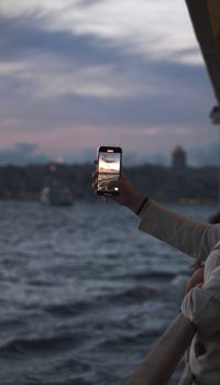 A person takes a photo of a scenic sunset over water using a smartphone from a balcony.