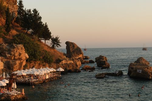 Free Calm seaside scene with rocky shoreline, trees, and boats at sunset. Stock Photo