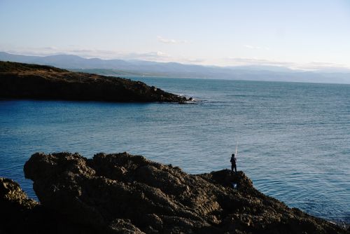 Free A lone fisherman enjoys a tranquil evening by the rocky seaside under a clear sky. Stock Photo