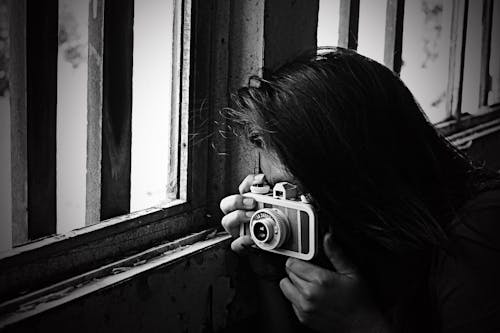 無料 窓の横で写真を撮る女性のグレースケール写真 写真素材