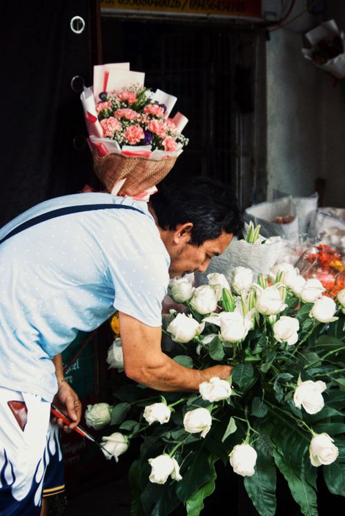 Free Florist carefully arranges white roses and holds a bouquet at a bustling market stall. Stock Photo