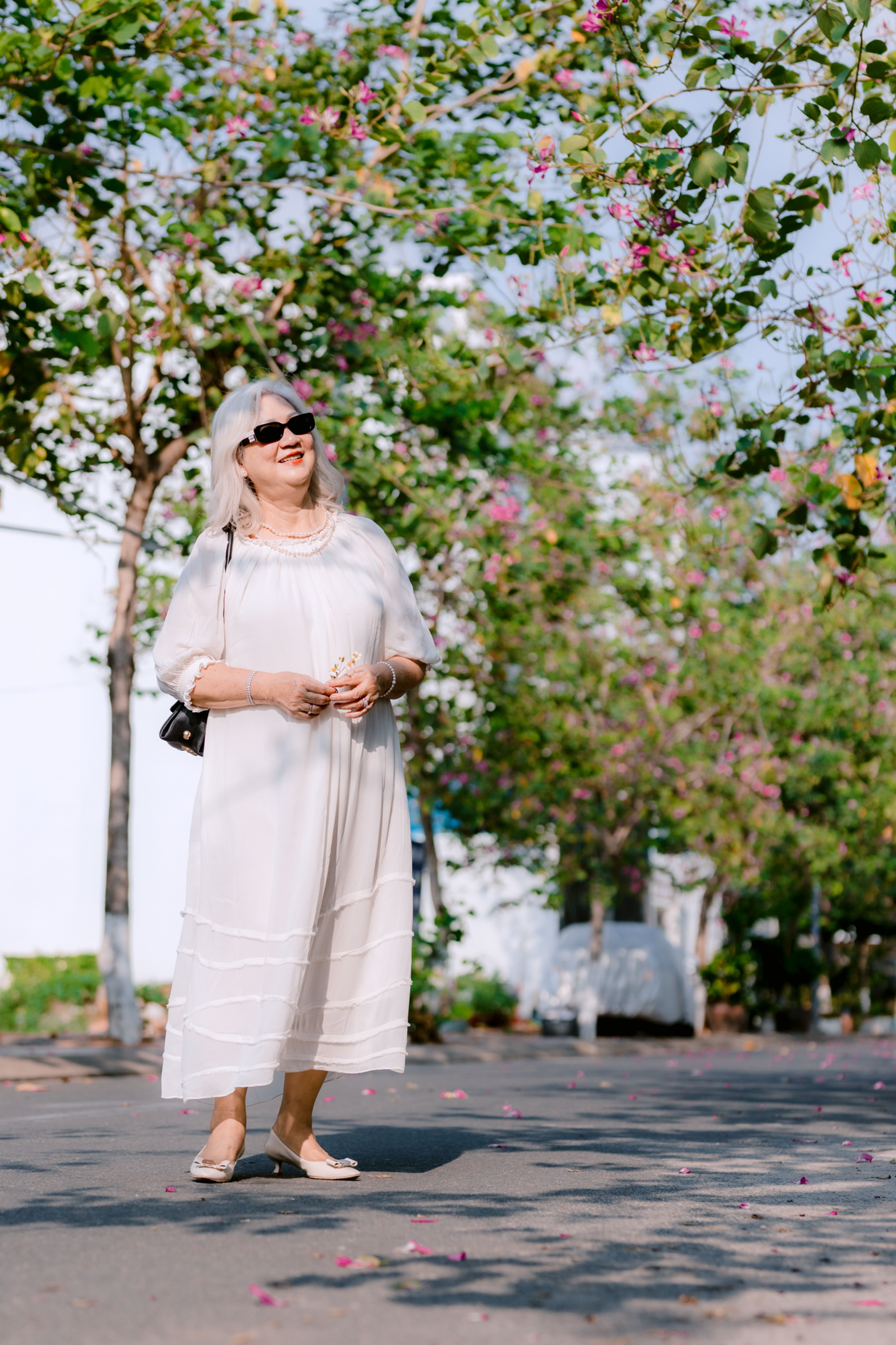 Stylish senior woman in white dress enjoys a sunny day outdoors surrounded by blooming trees.