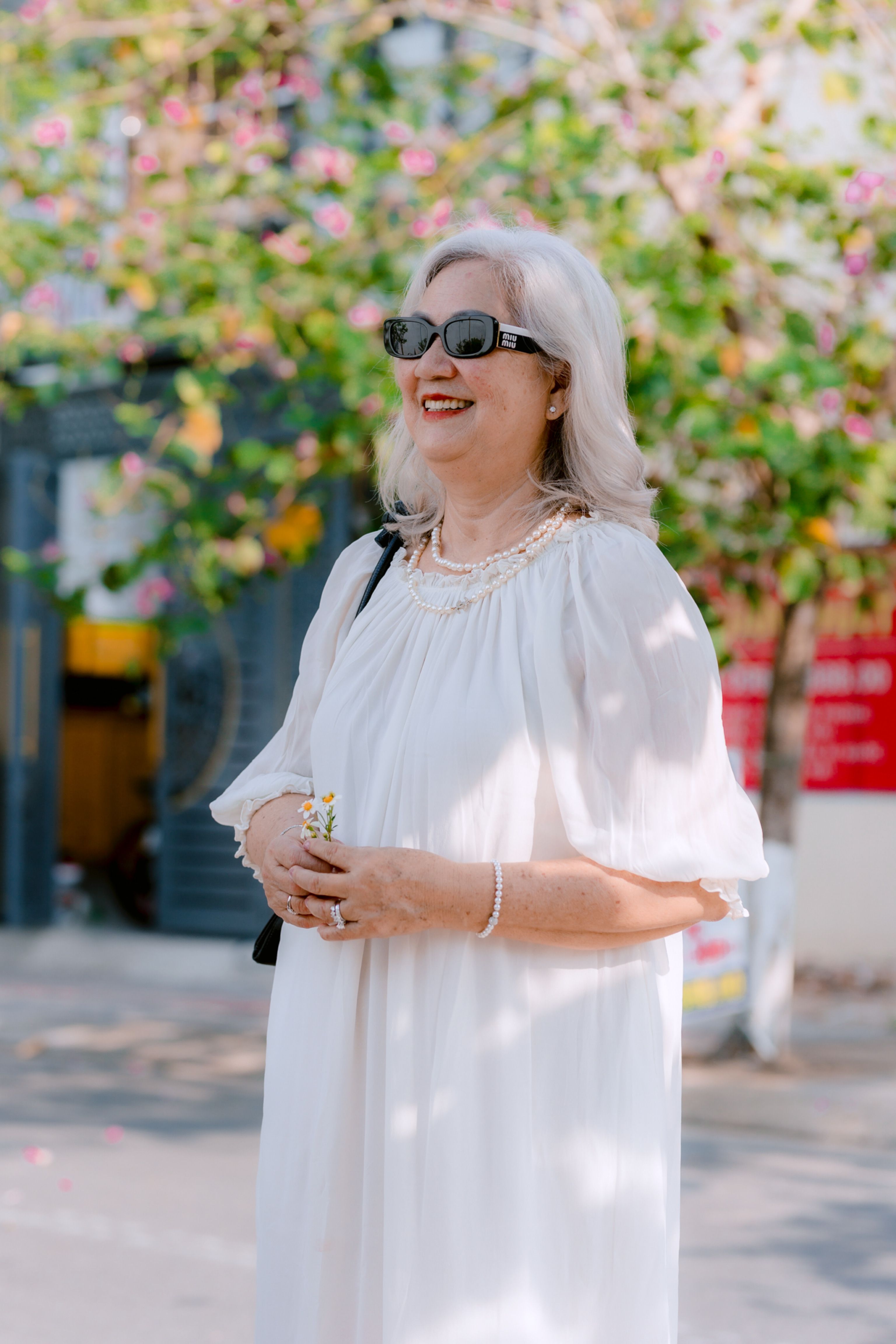 Smiling senior woman in white dress and sunglasses standing outdoors with flowers.