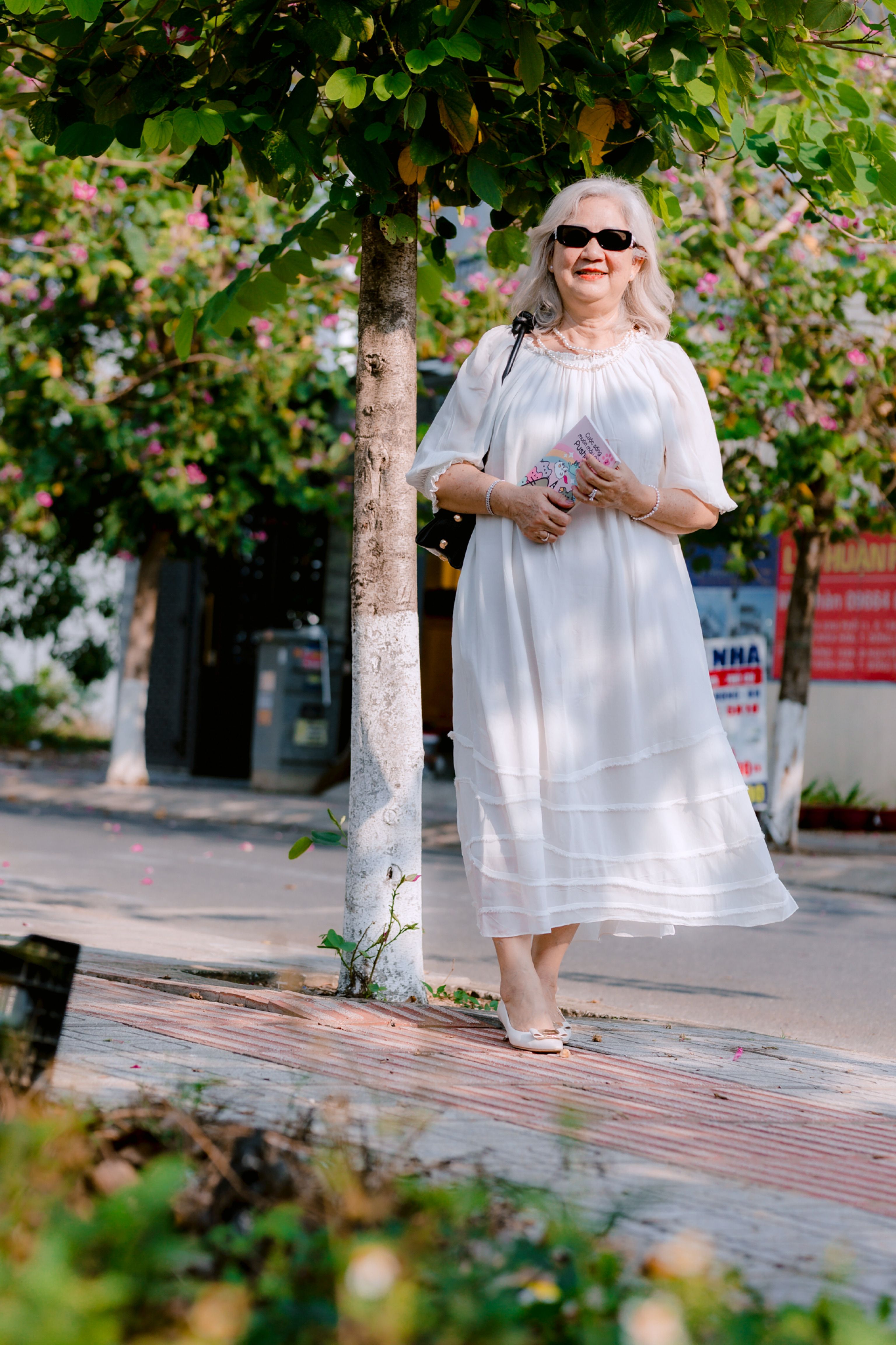 Stylish senior woman in white dress strolling confidently in a sunny park setting.
