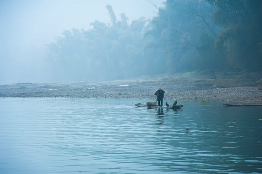 Misty morning on the river in Guilin, China. A lone person stands on a raft, surrounded by tranquil nature.