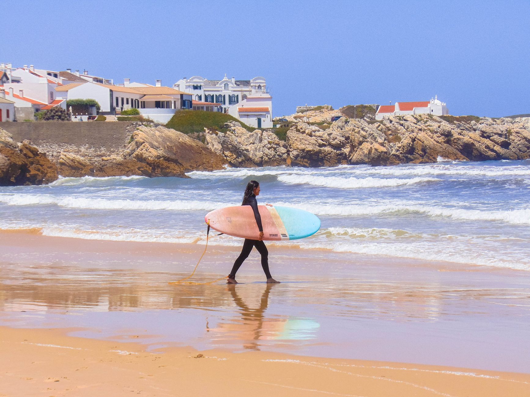 A surfer carrying a colorful surfboard strolls along the scenic beach in Peniche, Portugal.