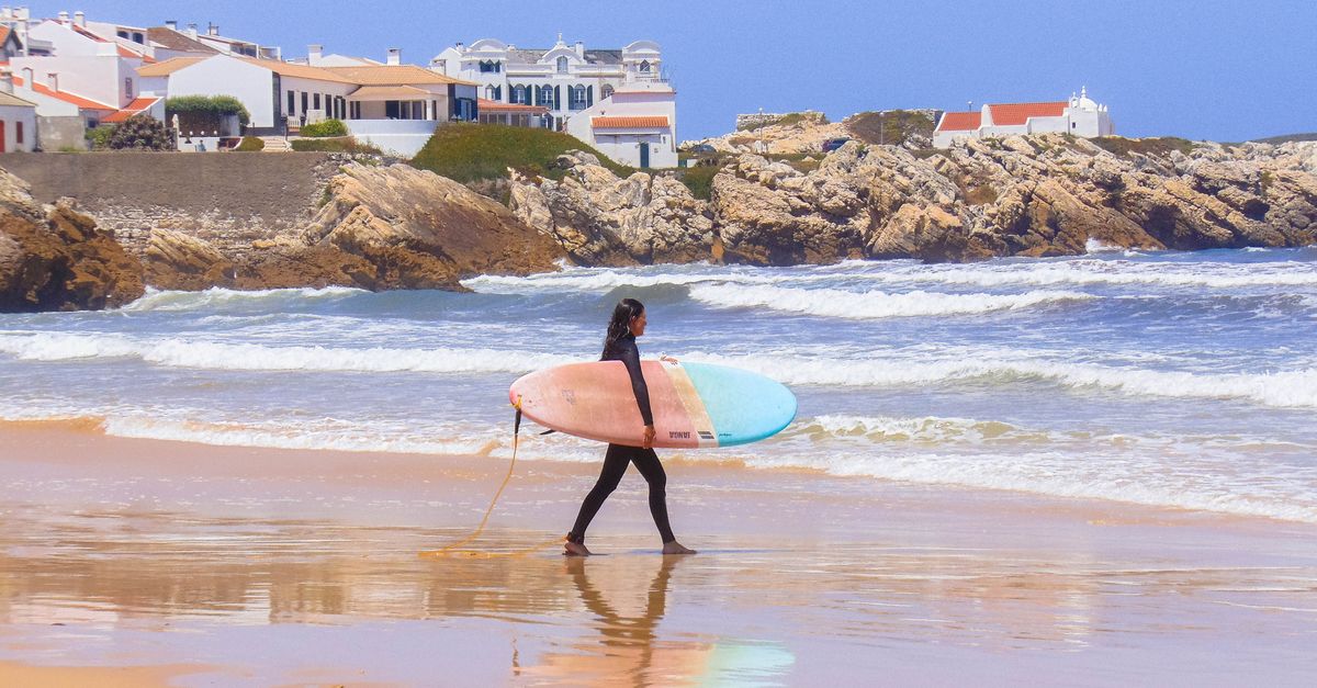A surfer carrying a colorful surfboard strolls along the scenic beach in Peniche, Portugal.