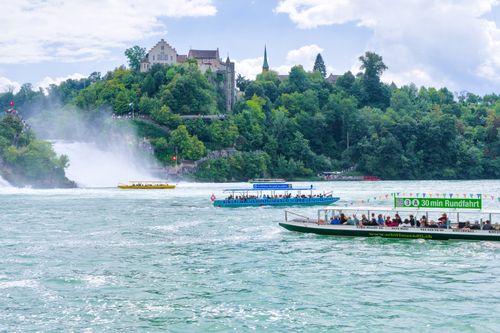 Free Tourists enjoy scenic views of Rhine Falls, Switzerland on boats. Lush greenery and castle enhance the scenery. Stock Photo