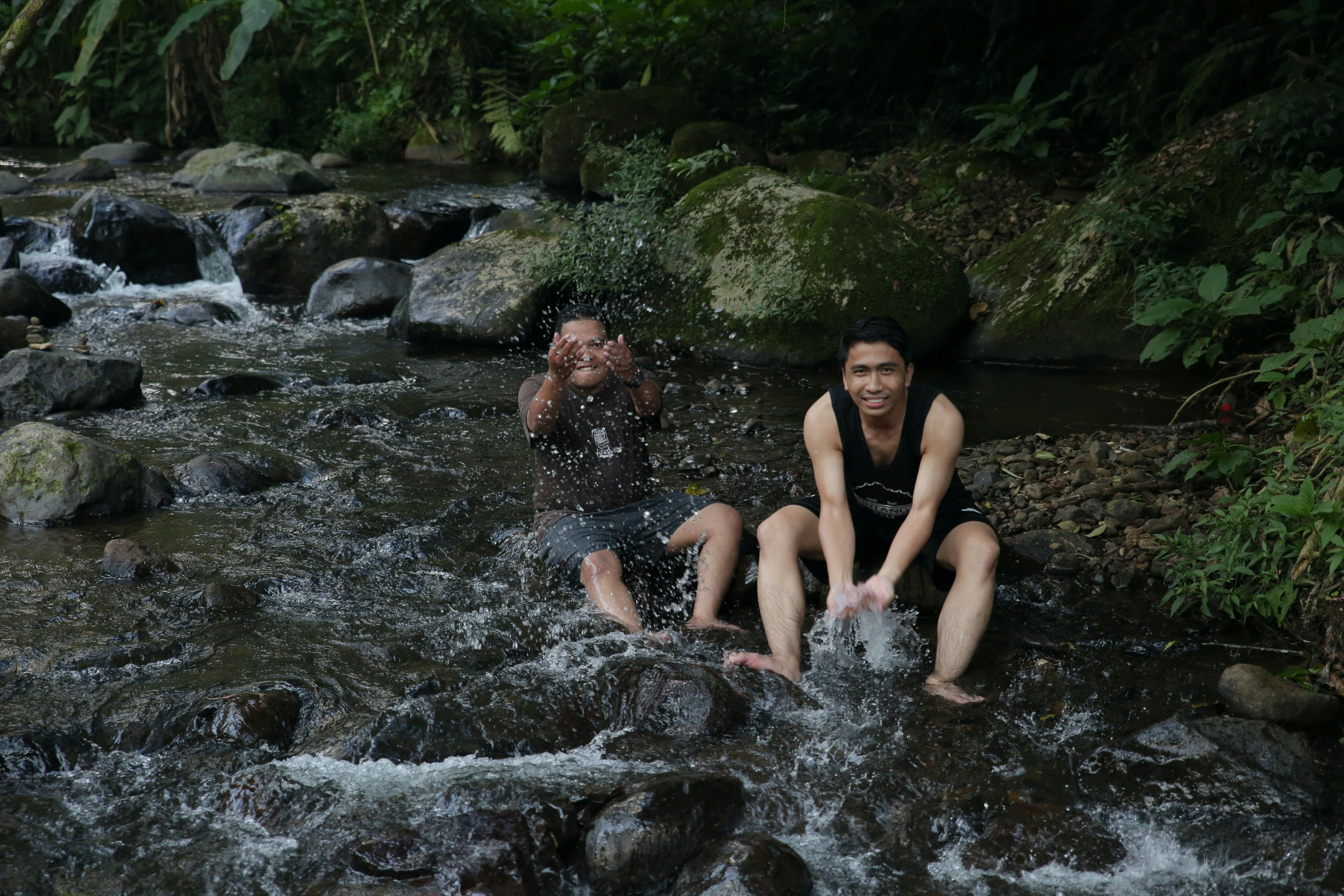 Two young men splash water playfully in a forest stream, enjoying nature.