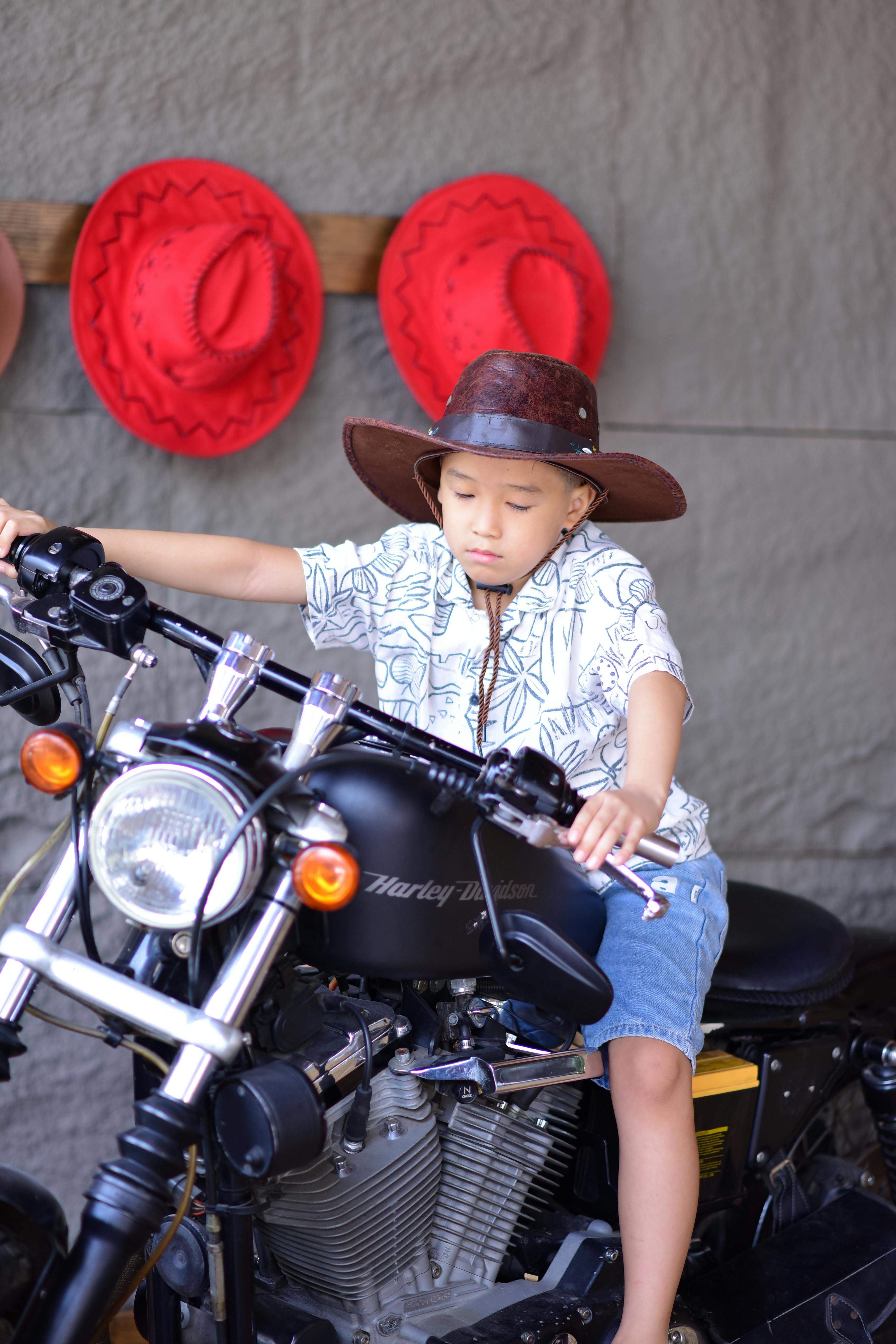 A boy wearing a cowboy hat sits on a motorcycle with red hats on display behind him.