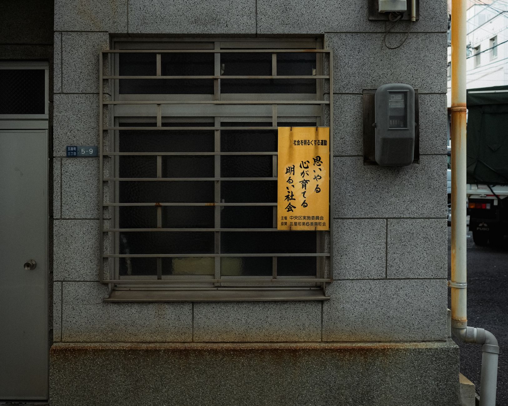 A Japanese urban building facade featuring a window with traditional signage and metal grating.
