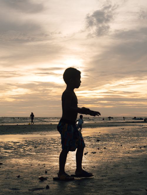 Free A serene beach scene in Thailand featuring a child's silhouette at sunset with the ocean in the background. Stock Photo