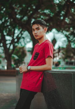 一名穿着红色衬衫的年轻男子在户外倚靠着一处表面，摆出时尚的姿势