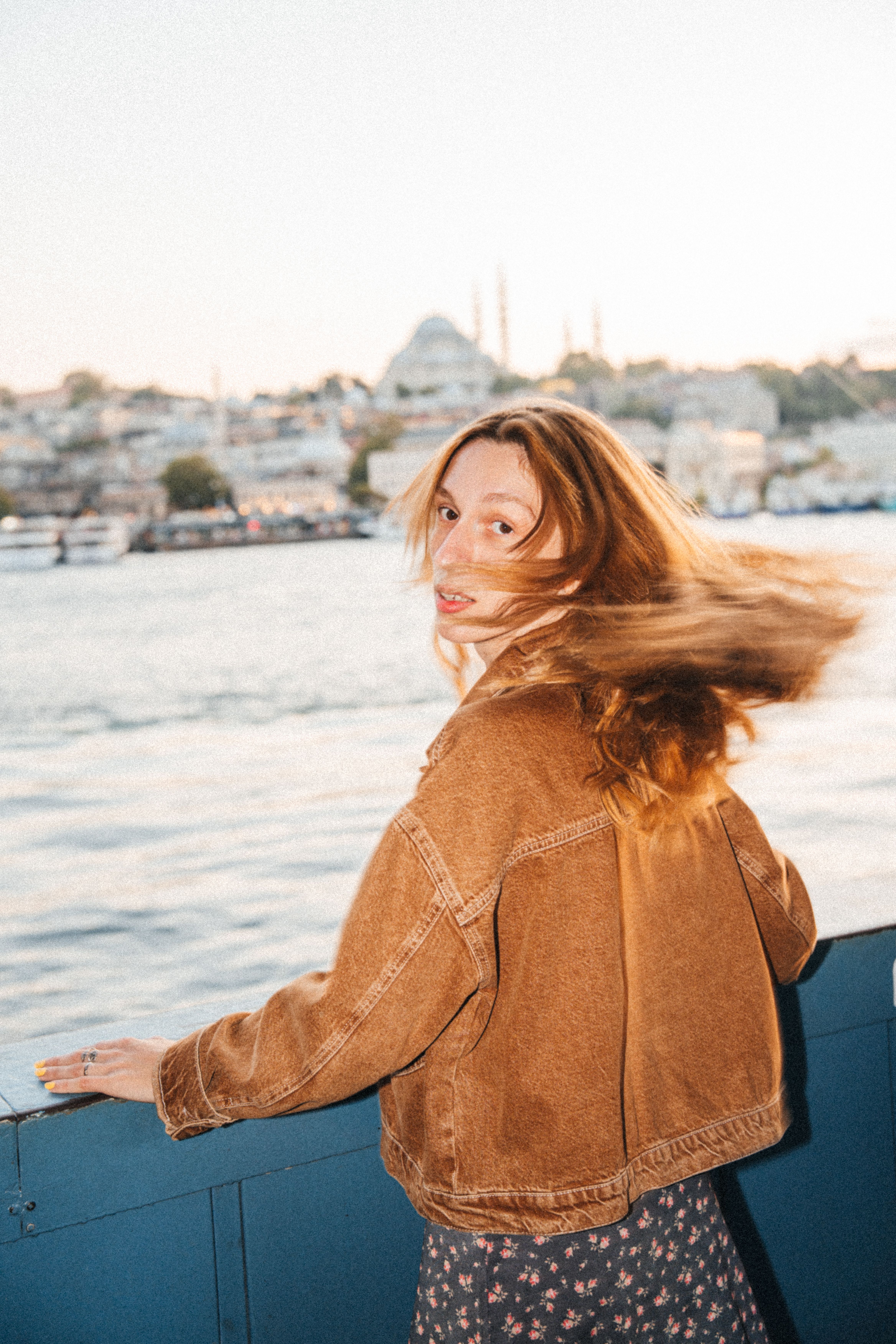 A young woman with flowing hair gazes towards the Bosphorus in Istanbul, with historic architecture in the background.