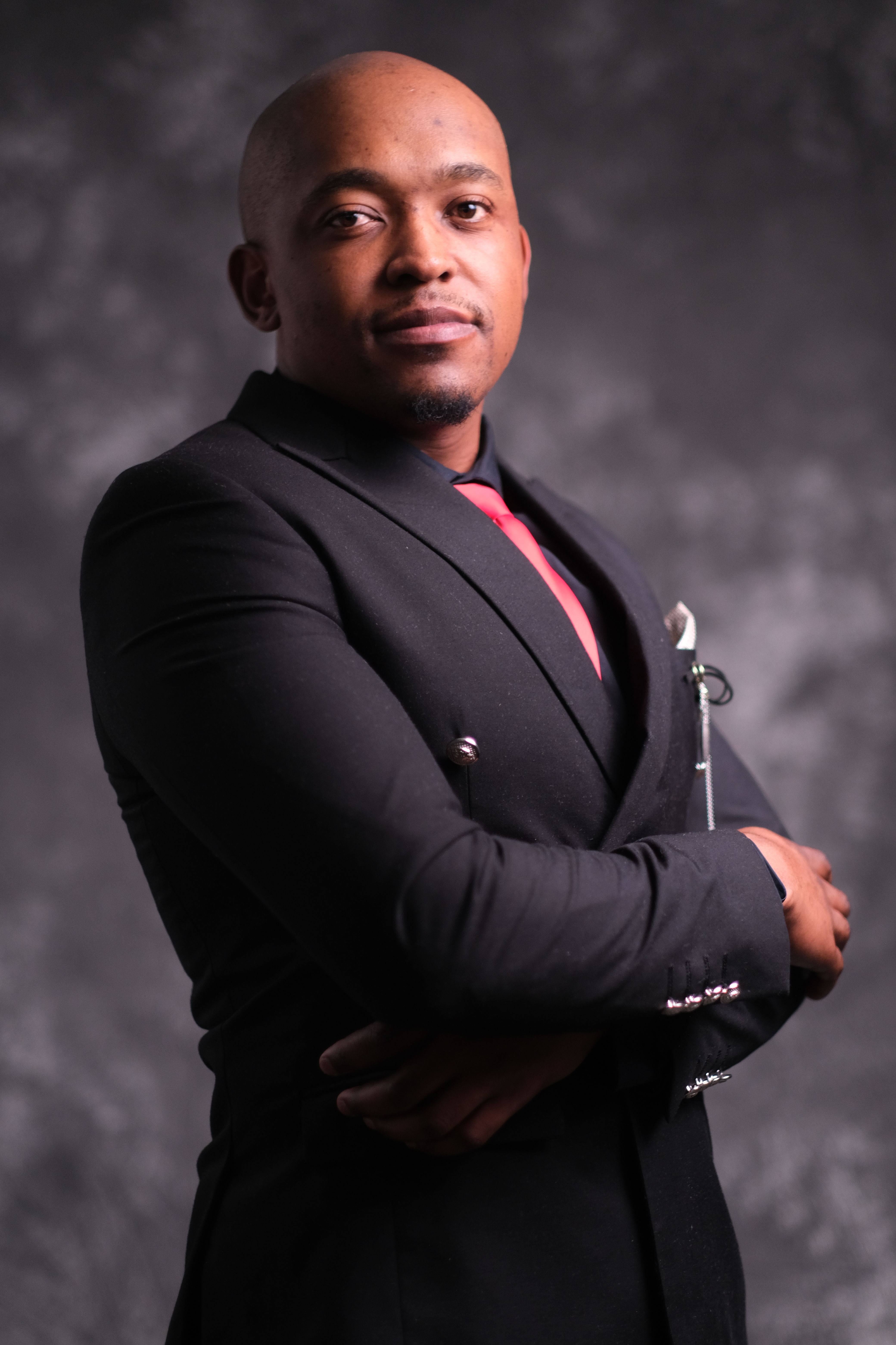 A confident businessman in a black suit poses with arms crossed against a textured background.