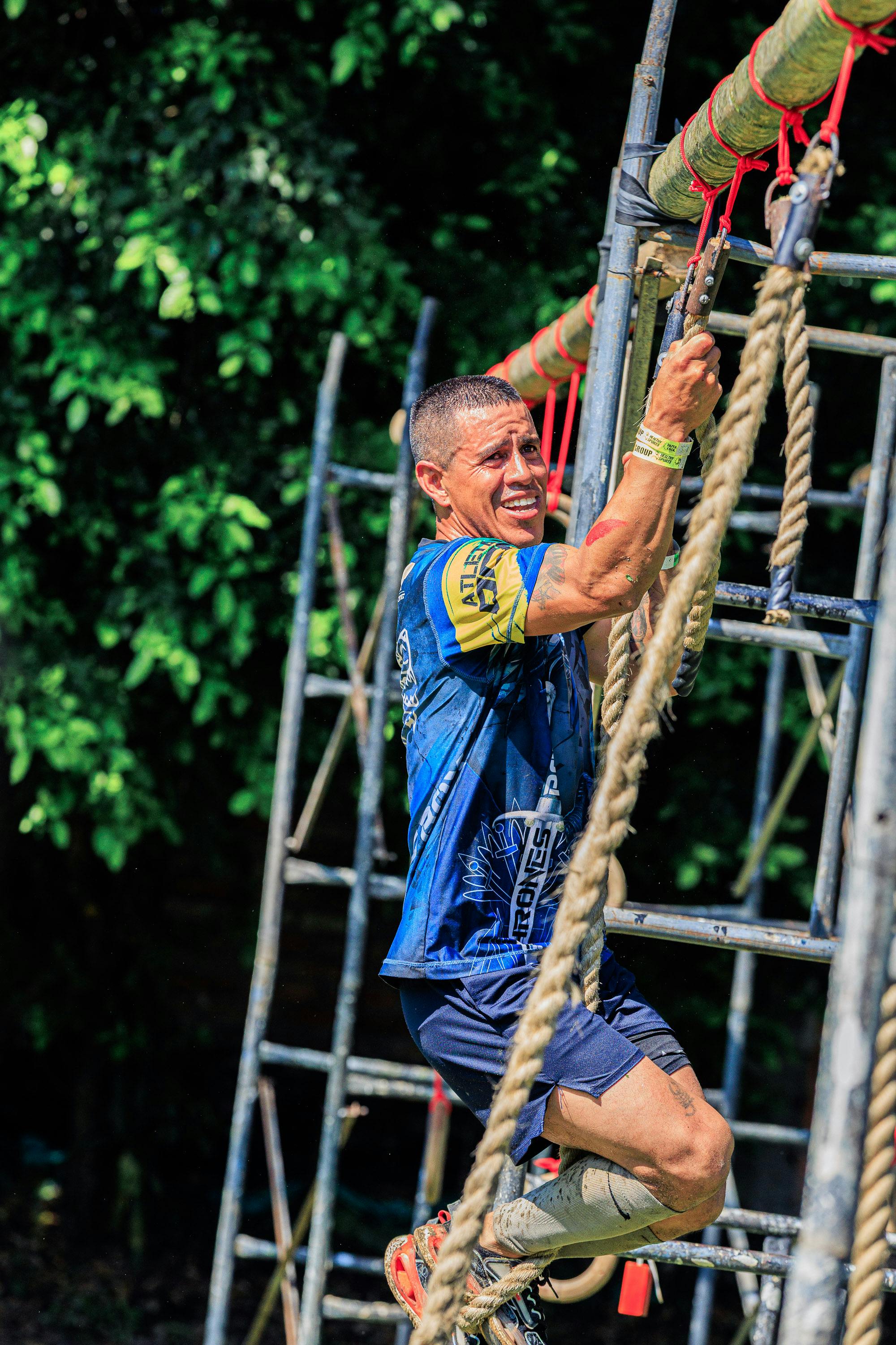 Athlete climbing a rope in an outdoor obstacle race on a sunny day.