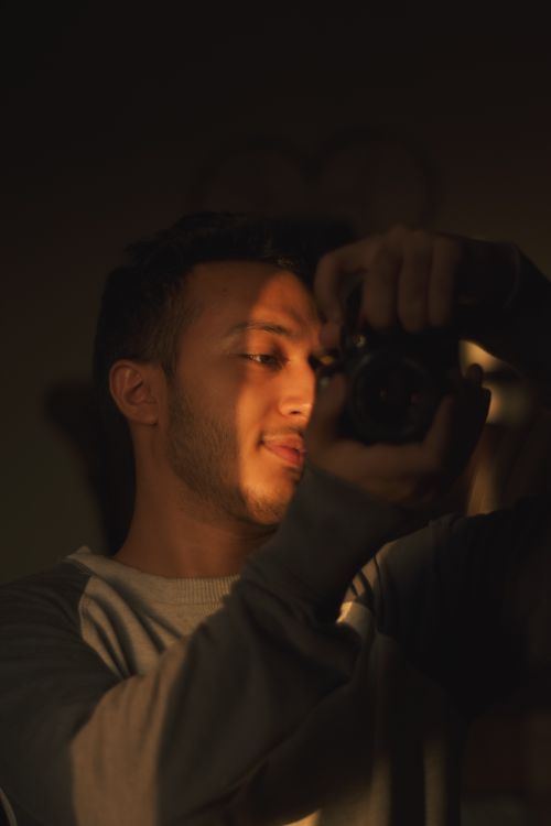 Free A candid portrait of a male photographer capturing a creative moment indoors, highlighted by warm lighting. Stock Photo