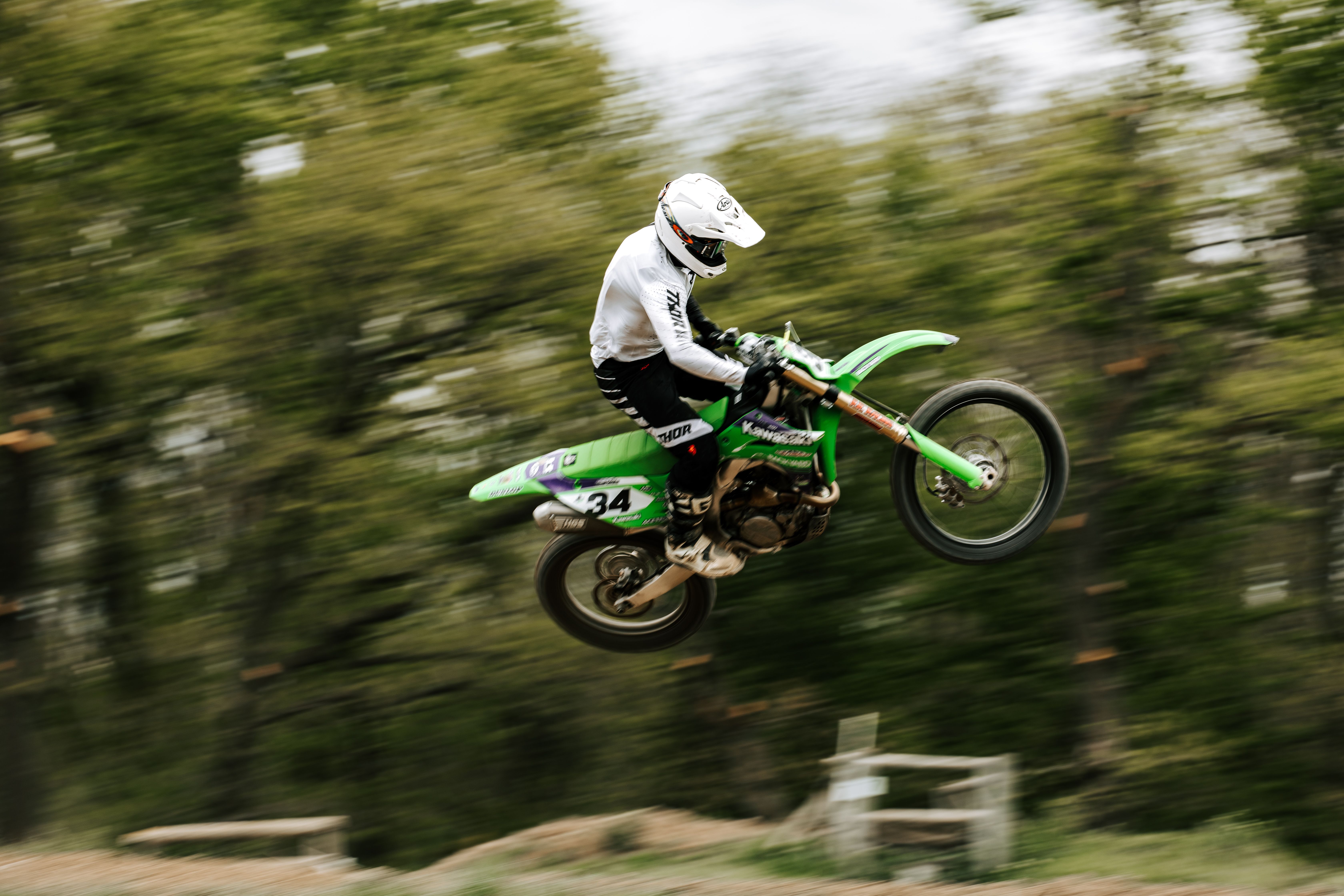 Dynamic capture of a motocross rider airborne on a green dirt bike with blurred forest background, showcasing speed and skill.