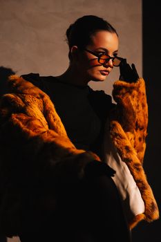 Portrait of a stylish woman wearing a luxurious fur coat and glasses against a dark background.
