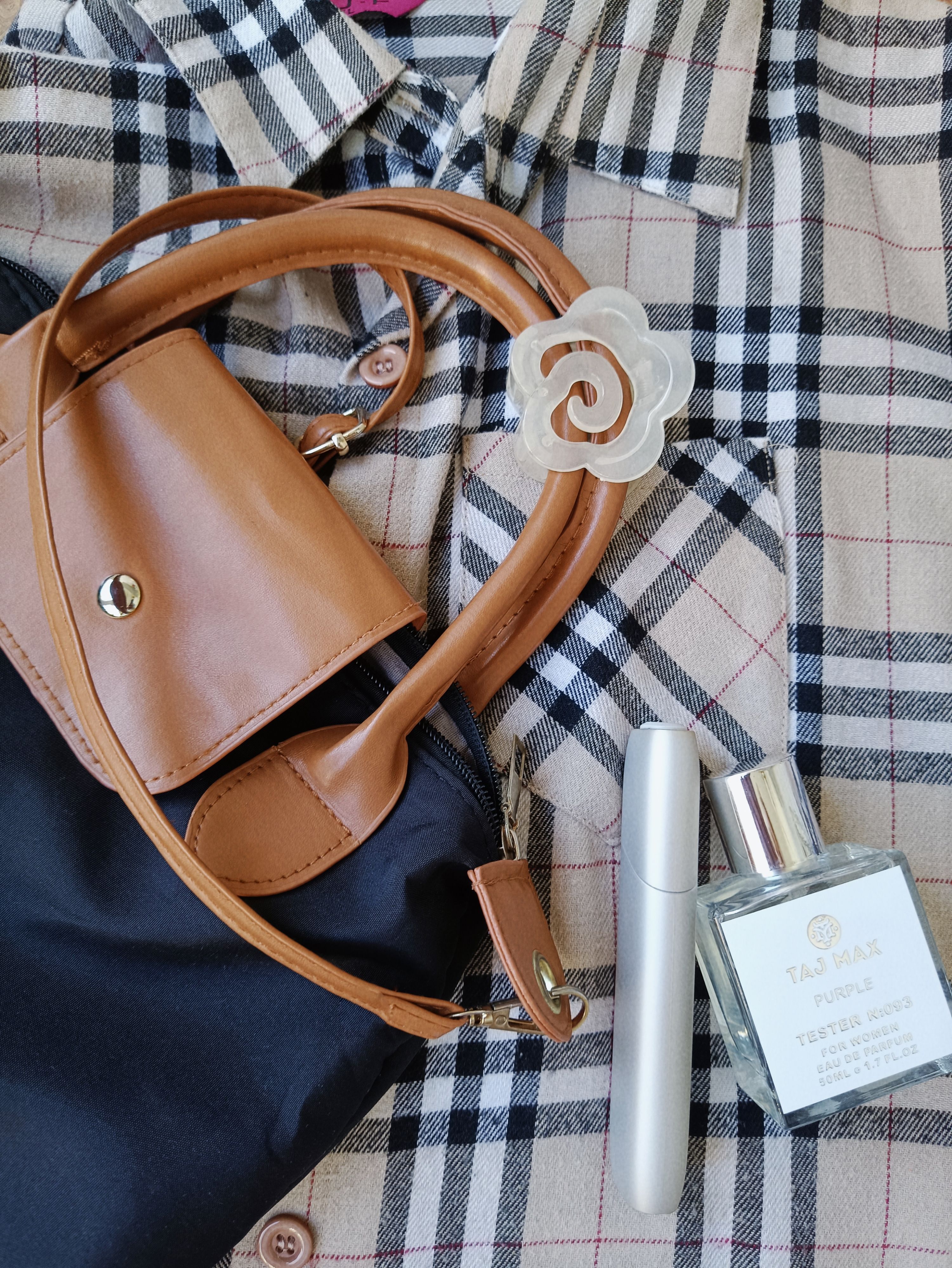 Flat lay de uma bolsa estilosa, perfume e acessórios sobre um fundo de camisa xadrez.