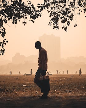 A silhouetted man walks through a tree-lined park as the city skyline looms in the hazy distance.