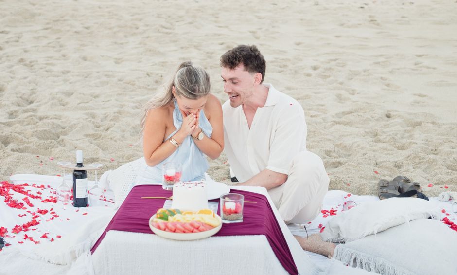 A couple enjoying a romantic picnic on the beach with a celebration cake and fruit.