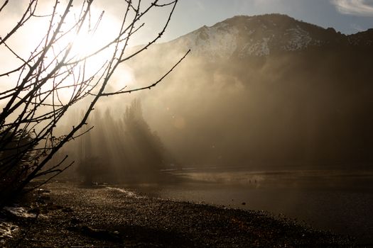 Sunlight streams through mist on a serene mountain landscape near a lake during winter.