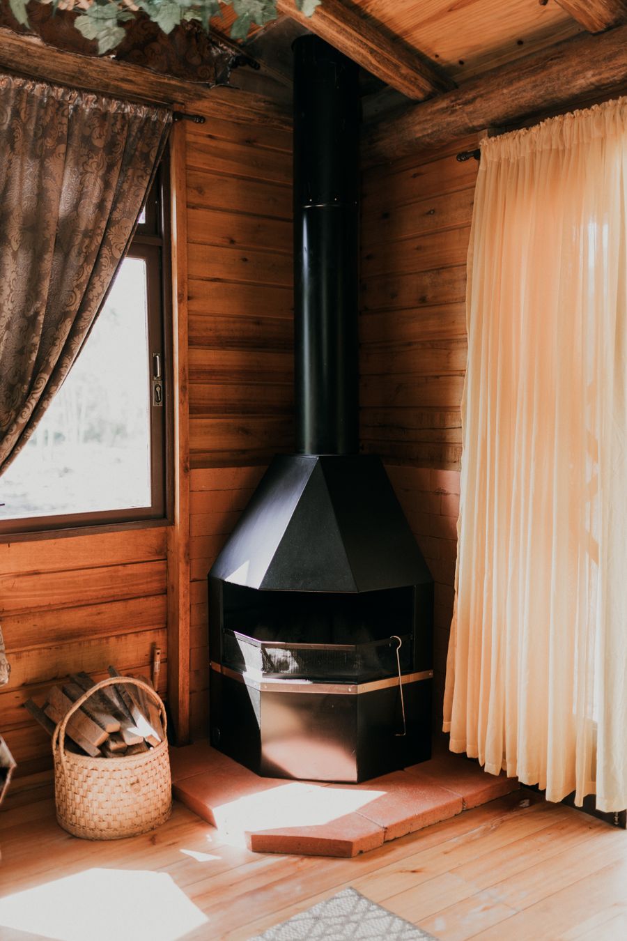 A black wood stove on a raised hearth in a pine-panelled cabin room, a basket of logs beside it