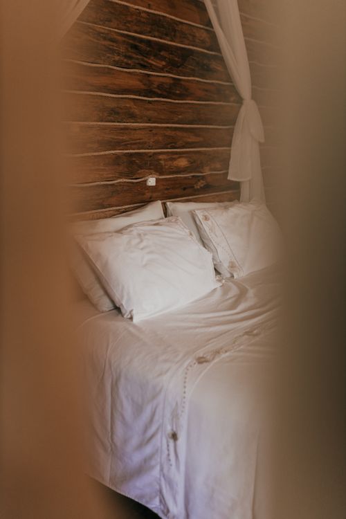 Free A rustic bedroom interior featuring a comfortable bed with white linens against a wooden wall, framed by soft curtains. Stock Photo