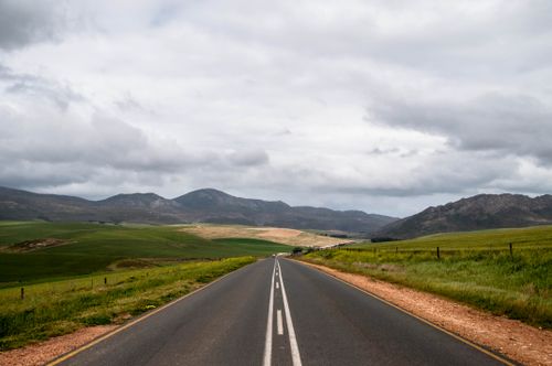Free A picturesque road in South Africa surrounded by vast fields and mountains under a cloudy sky. Stock Photo