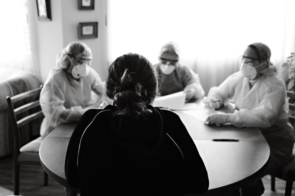 Healthcare workers in protective gear discuss with a patient during a medical consultation in a home setting.