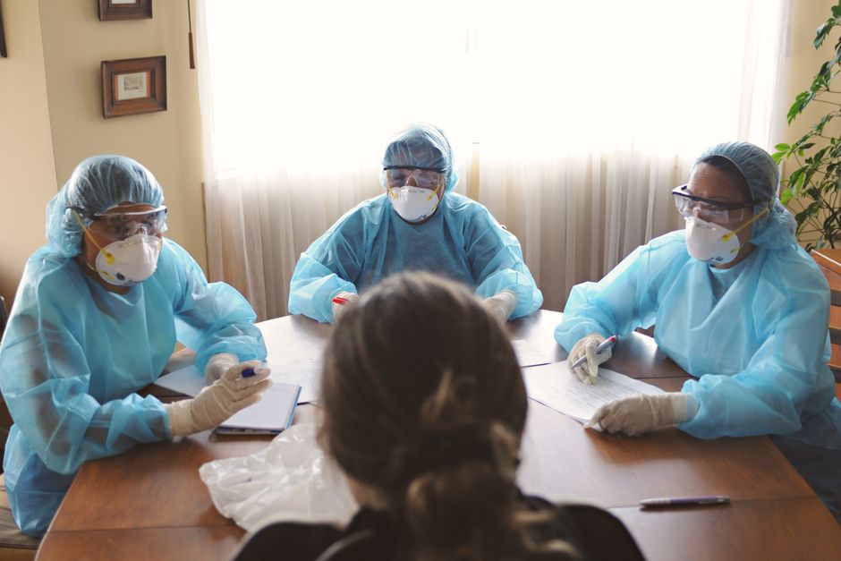 Grupo de trabajadores de la salud en EPI manteniendo una discusión alrededor de una mesa en un interior.
