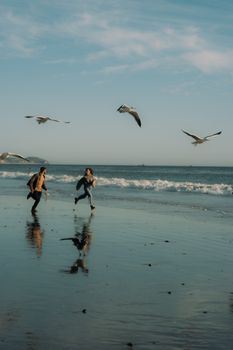 A joyful couple runs along a sunny Los Angeles beach, surrounded by seagulls.