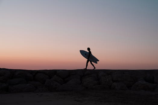 Silhouette of a surfer walking with a surfboard at sunset on a rocky beach in Portugal.