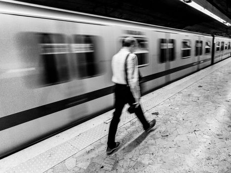 Black and white image of a person walking in a subway station with a moving train.