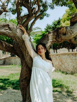 A woman in a white dress poses gracefully beside a large tree in a sunlit park setting.