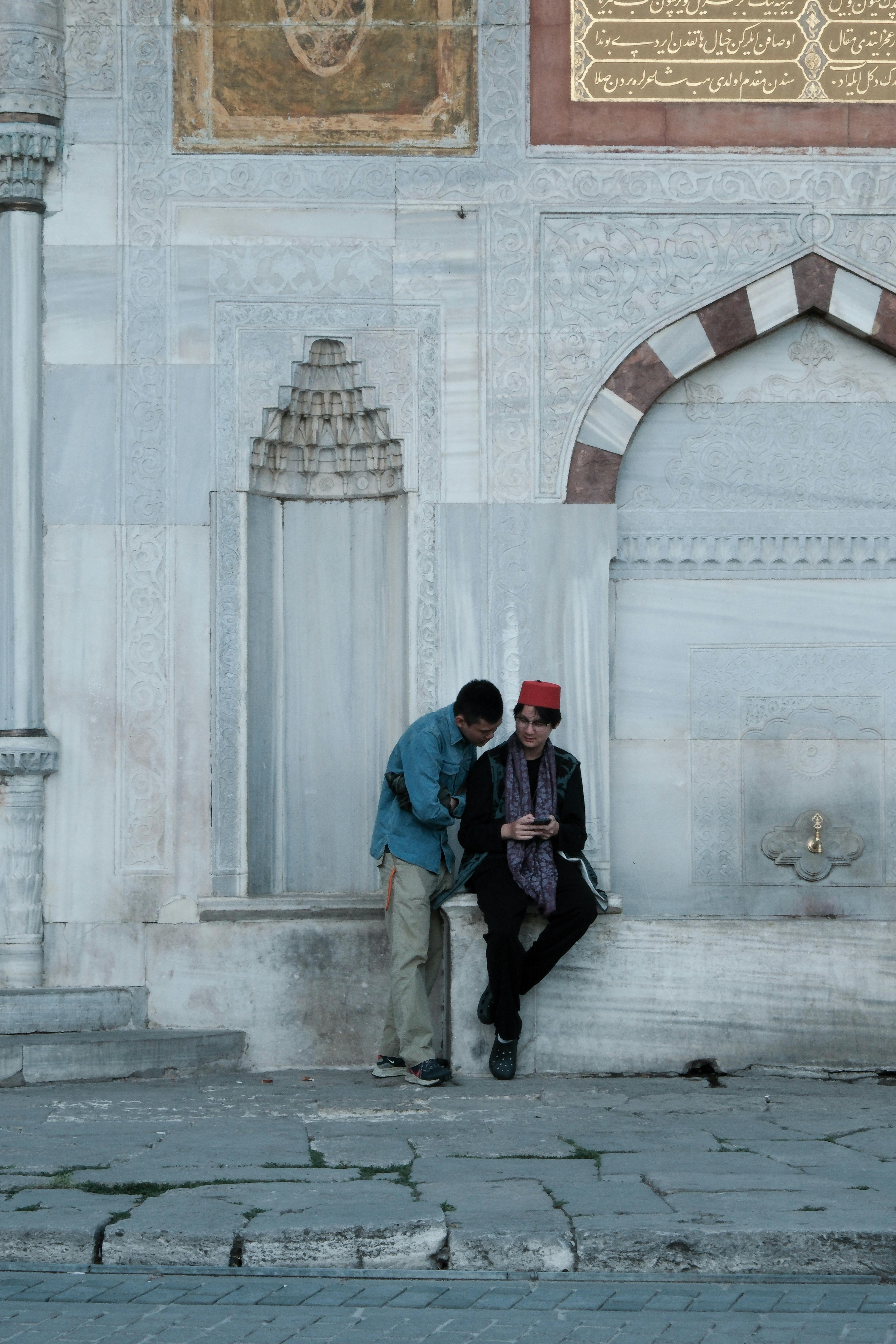 Two men by Ottoman-style wall in casual conversation, showcasing cultural attire and historic architecture.