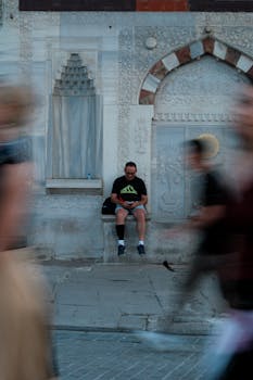A man sitting against a historic wall while people walk by in motion blur.