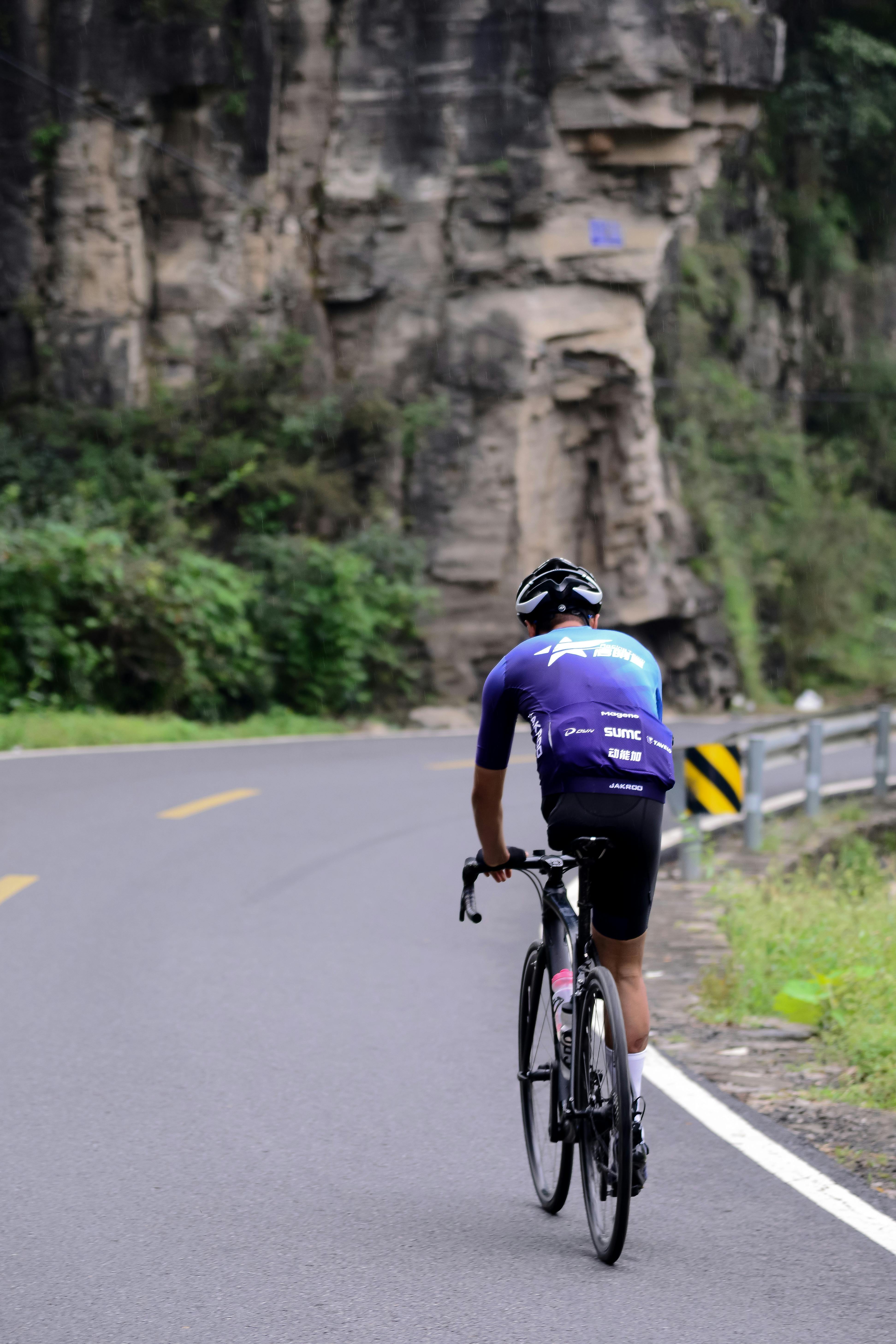 A cyclist in sports gear rides along a winding mountain road surrounded by cliffs and greenery.