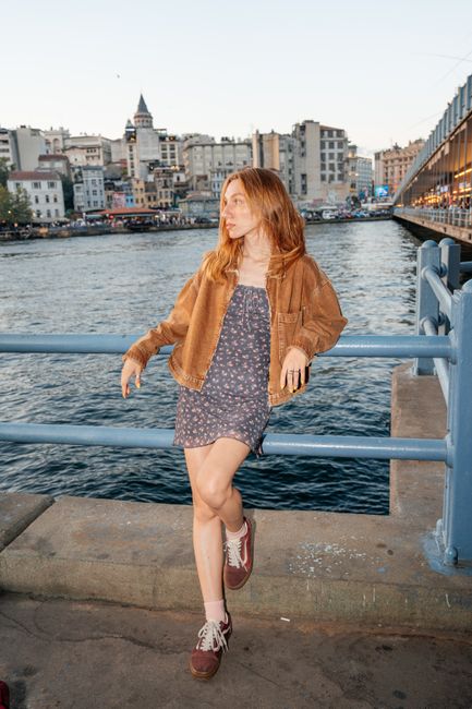 A young woman in a casual outfit poses by the Galata Bridge in Istanbul, with a cityscape backdrop.